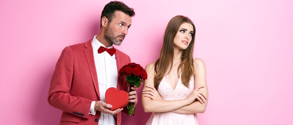 A couple against a pink wall looking annoyed holding flowers &candy