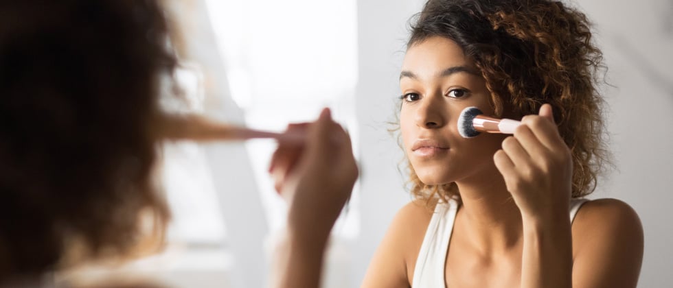 Young woman looking into the mirror while brushing on makeup