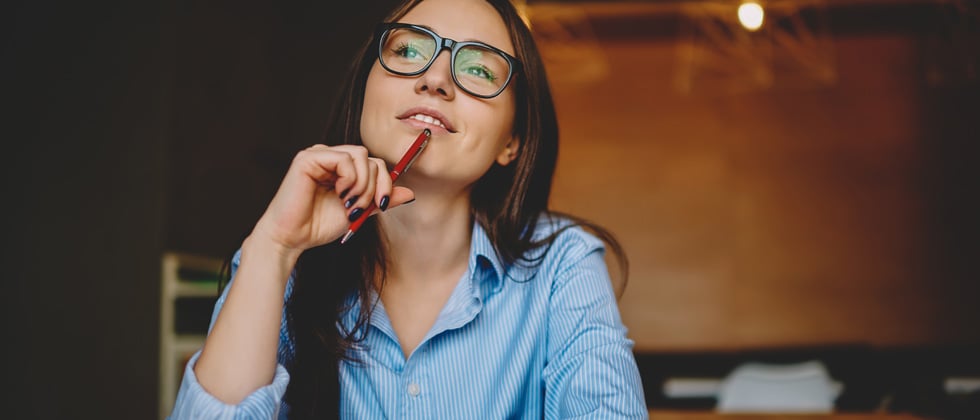 A woman with glasses sitting with a pen to her chin thinking