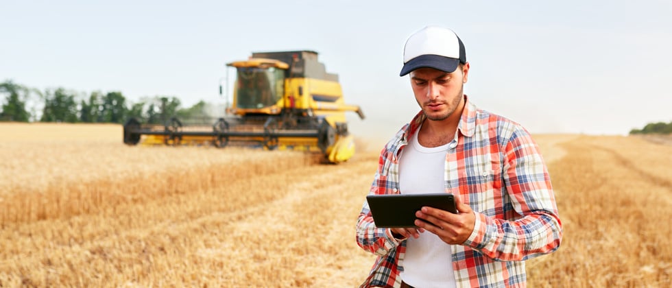 A male farmer holding a tablet in a field with a tractor behind him