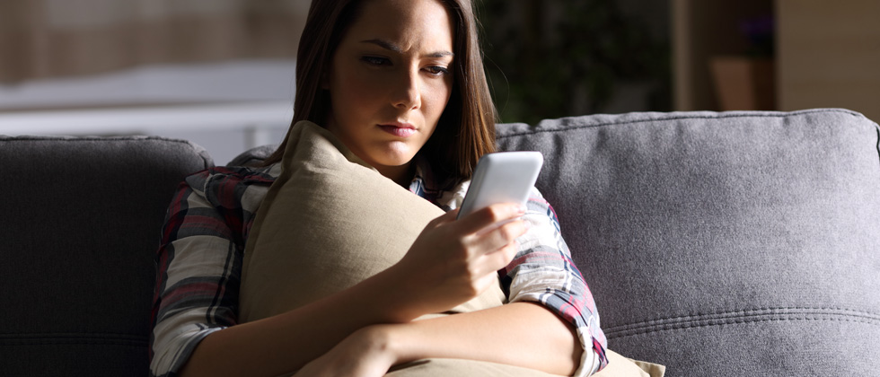 A woman sitting and holding a pillow while looking at her phone sadly