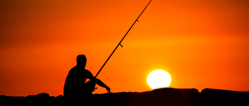 A man sitting next to a body of water fishing at sunset