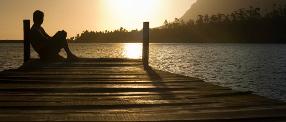 A man sitting at the edge of the dock reflecting at sunset