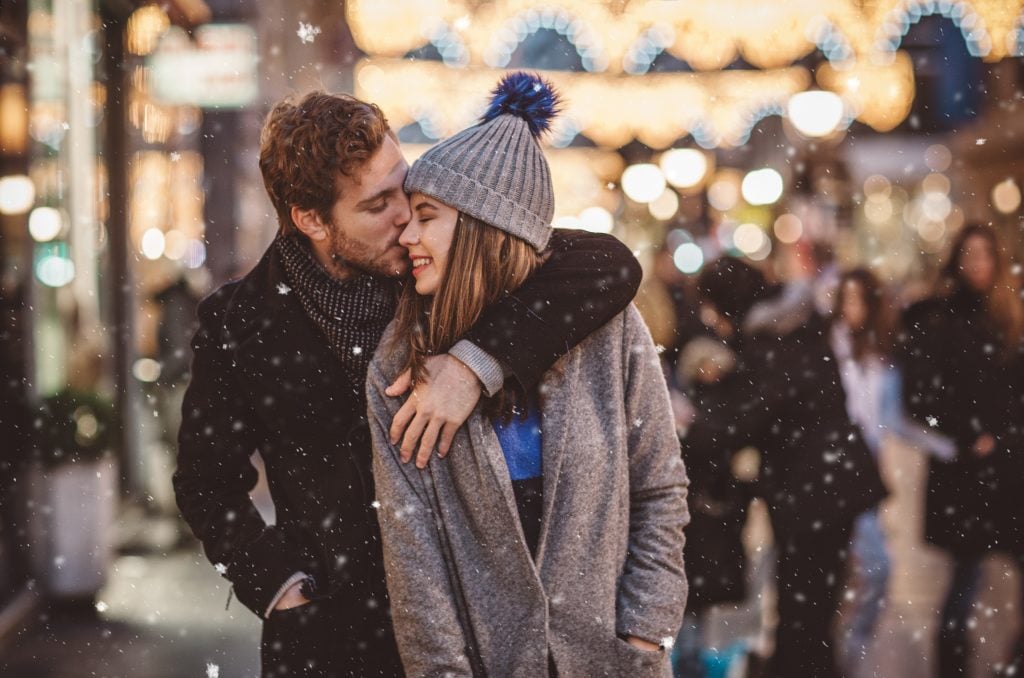 Couple enjoying a snowy christmas in the city while he is kissing her cheek.