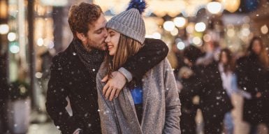 Couple enjoying a snowy christmas in the city while he is kissing her cheek.