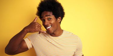 A young man stands against a yellow background, making a 'phone me' gesture with his right hand