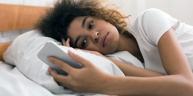 A young woman, looking troubled, lies in bed staring at her phone