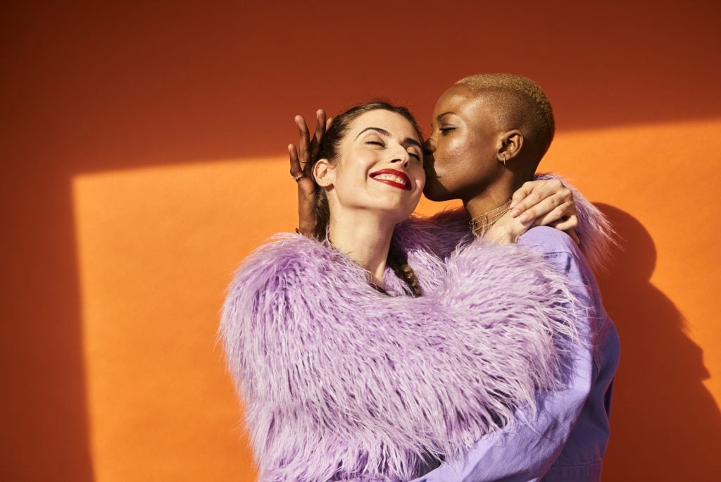 Two women having fun and one kissing the other on her cheek. Both wearing violet fashion pieces.