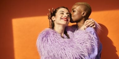 Two women having fun and one kissing the other on her cheek. Both wearing violet fashion pieces.