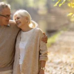 A couple walk side by side along a gravel path near a forest in the sunlight.