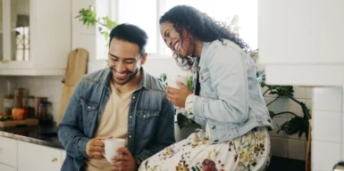 A couple sitting on the kitchen corner holding cups and laughing.