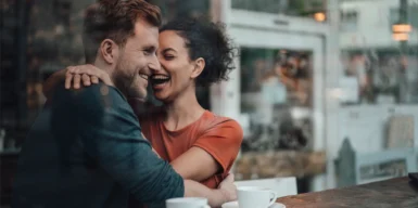 A couple in their 30s laughing and hugging in a coffeeshop