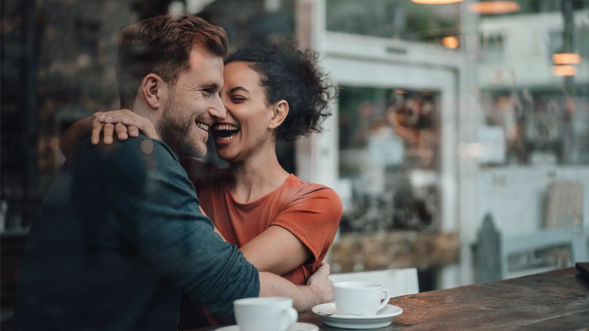 A couple in their 30s laughing and hugging in a coffeeshop