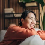 Asian Women on the couch in her living room looking out of the window.